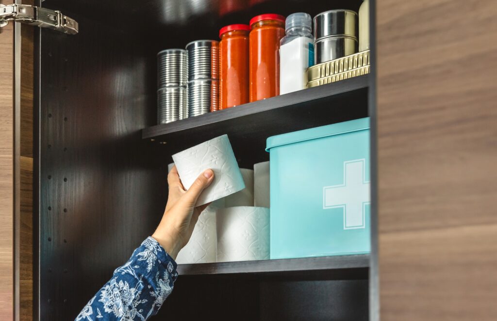 Close up of unrecognizable female hand filling toilet paper to emergency food pantry in kitchen, preparing for potential disasters. Alert, foresight and self sufficiency. 72 hours survival kit concept